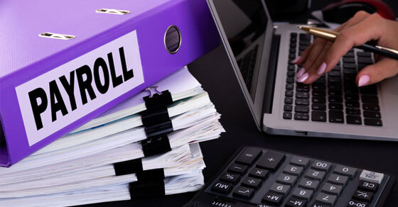 Binder labeled "payroll" on top of stacks of paper sitting next to a keyboard with a persons hand on top of the keys.