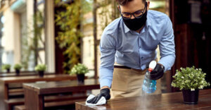 employee wiping down a table in a restaurant wearing gloves and a mask.