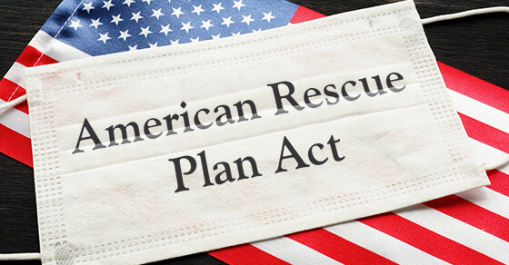 disposable mask on top of the american flag. the mask has the words "American Rescue Plan Act"