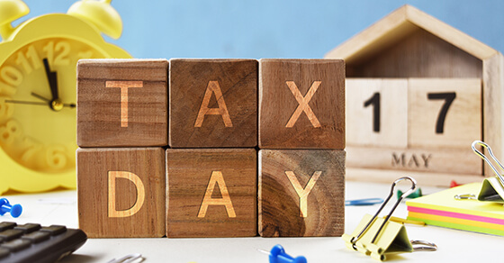 table with block letters spelling out "Tax Day" on it. Behind the blocks is a clock and a calendar with the date May 17