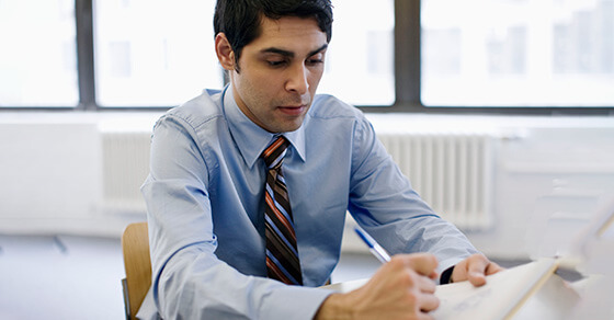 Individual in a room, sitting at a table, looking at a document with a pen in hand.