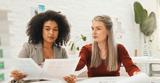 Two businesspeople collaborate on a project together. Team of designers reading documents together. Mature businesswoman working with her staff member.