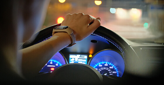 Close up of driver hands holding steering wheel driving car with blurred city street lights on background at night.