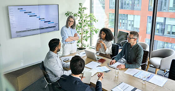 Senior female ceo and multicultural business people discussing company presentation at boardroom table. Diverse corporate team working together in modern meeting room office. Top view through glass