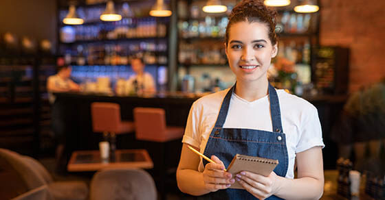 Young smiling waitress in workwear standing in front of camera in luxurious restaurant and going to writie down order of client