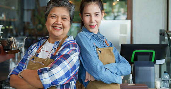 Senior mother with Asian daughter, standing in front checkout counter taking cafe, which is its own bakery, stood with their apron pockets in their pockets, raising their hands make hearts.