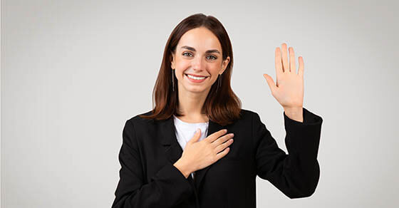 Pleasant millennial caucasian businesswoman with a sincere smile raises her right hand to take an oath or pledge, placing her left hand over her heart, signifying honesty and commitment