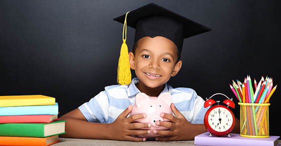 Young African American school boy sitting at desk with books, pencils and piggybank on black background