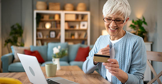 Smiling elderly woman is holding credit card and using laptop. Sitting at dining table at home. Making online payment. Purchasing goods or services on internet. Enjoying benefits of modern technology