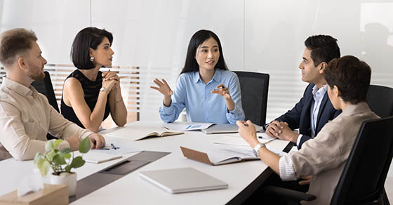 Confident young speaker woman talking to multiethnic team. Female boss, leader discussing teamwork strategy with employees. Group of stakeholders, partners meeting at large table
