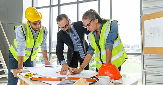 Construction engineer and architect in vest and helmet discussing blueprint with construction manager, standing on construction site