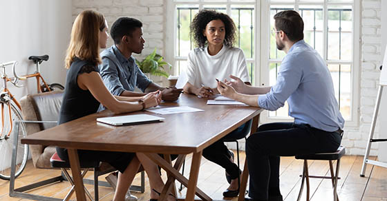 Full length motivated young multiracial business people sitting at table creating project ideas discussing problem solution negotiating working issues or brainstorming at group office meeting.