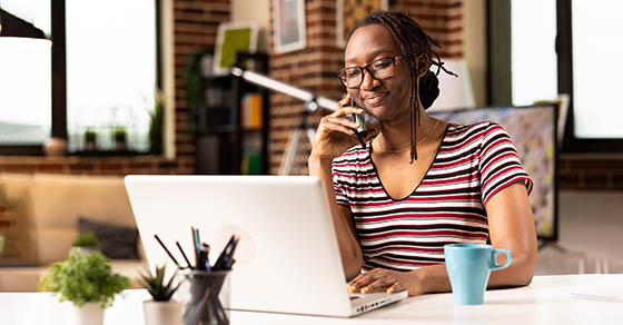 Self employed woman sits at desk, multitasking, speaks with clients on phone and updates data on laptop. Smiling female freelancer uses mobile device while working from home on personal computer.